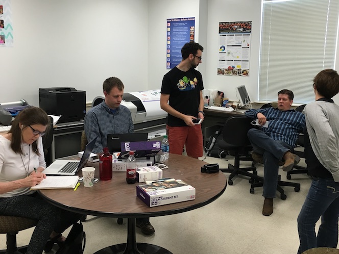 team members working in the Media Lab: Leslie and Andrew are at their laptops on a table, Jack is standing holding his phone, Rick sits leaning back in a chair; Jamie is in the forefront with her back to the camera