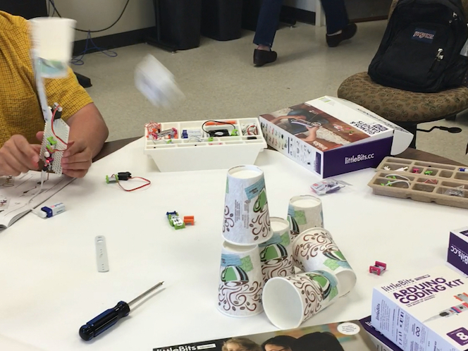 a man on the left holds the littleBits throwing arm invention, launching a paper ball toward a tower of plastic cups, with some of the cups already knocked over