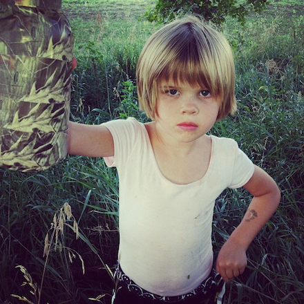 A little girl with a dirty blonde bob haircut fills much of the frame, holding a camoflauged container the size of mason jar up and out with her right hand. The look on her face is stoic.