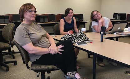 Joyce sits in the foreground, looking up toward an off-image television; Sarah and Erica look toward the camera, smiling