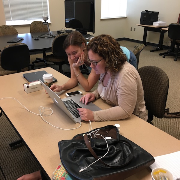 Erica sits at a table in a classroom working on her MacBook Pro; Sarah leans over her right shoulder, smiling at the laptop screen