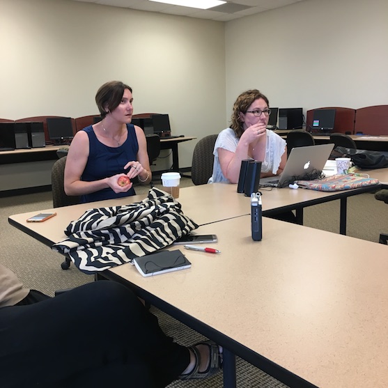 Sarah and Erica sitting at a table looking up and forward; Erica has her laptop in front of her; Sarah is holding an apple