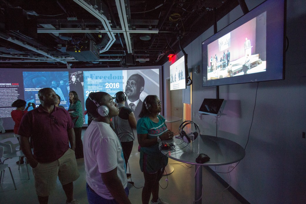 Banner picture showing event attendees listening to the speech using headphones while they roam around a room with walls covered with pictures of the historic event